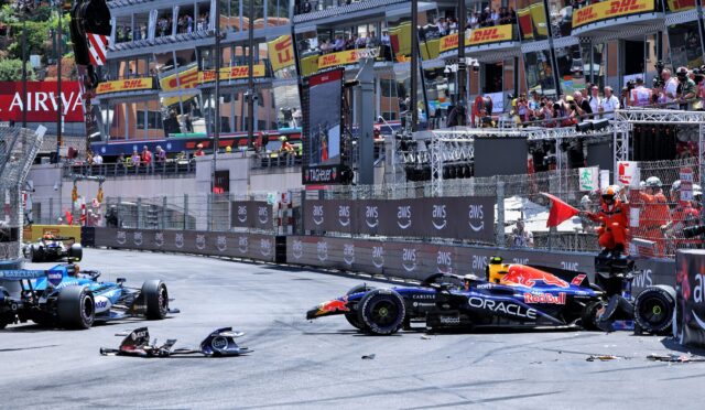 Formula 1 cars stop on a street circuit after a crash, with debris and a damaged front wing on the track and spectators watching from the barriers and grandstands behind safety fencing.