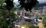 Framed view of Monaco city harbor race circuit with grandstands and boats, greenery in the foreground.