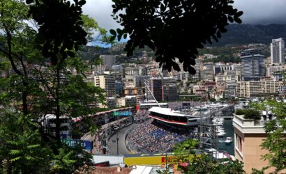 Framed view of Monaco city harbor race circuit with grandstands and boats, greenery in the foreground.