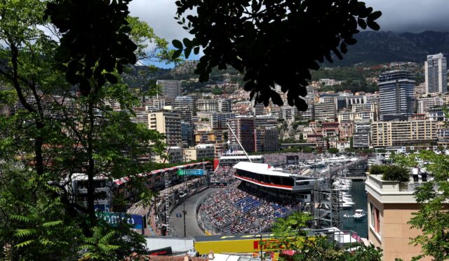 Framed view of Monaco city harbor race circuit with grandstands and boats, greenery in the foreground.
