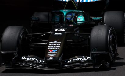 Front view of a teal-and-black Formula 1 car with sponsor logos, in a dark pit area, nose and cockpit visible.