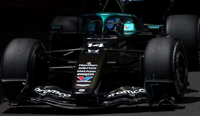Front view of a teal-and-black Formula 1 car with sponsor logos, in a dark pit area, nose and cockpit visible.