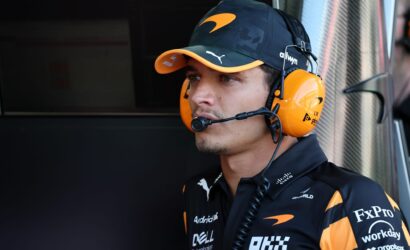 Pit crew member wearing an orange headset and cap, focused in a workshop-like setting with sponsor logos on dark uniform.