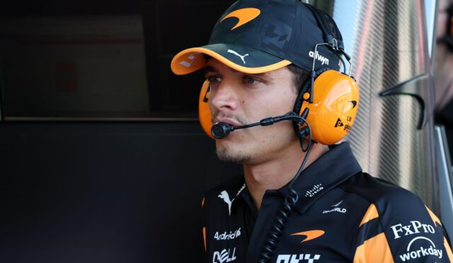 Pit crew member wearing an orange headset and cap, focused in a workshop-like setting with sponsor logos on dark uniform.