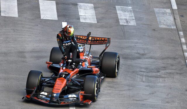 Top-down view of an orange Formula 1 car with a helmeted driver navigating a street circuit corner on a gray track with white markings.