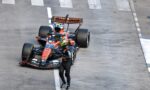 Top-down view of an orange and blue Formula 1 car parked on a race track, with a helmeted driver standing nearby on the white-lined tarmac.