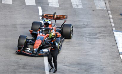 Top-down view of an orange and blue Formula 1 car parked on a race track, with a helmeted driver standing nearby on the white-lined tarmac.