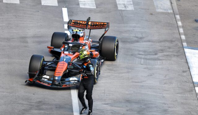 Top-down view of an orange and blue Formula 1 car parked on a race track, with a helmeted driver standing nearby on the white-lined tarmac.