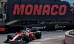 Red Formula 1 car on a tight Monaco street circuit, with a large Monaco sign on the barrier in the background.