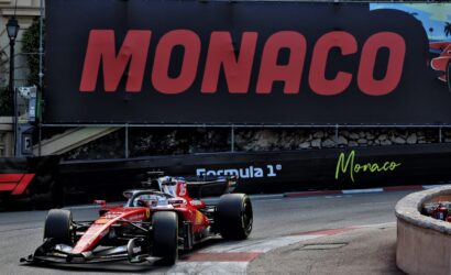 Red Formula 1 car on a tight Monaco street circuit, with a large Monaco sign on the barrier in the background.
