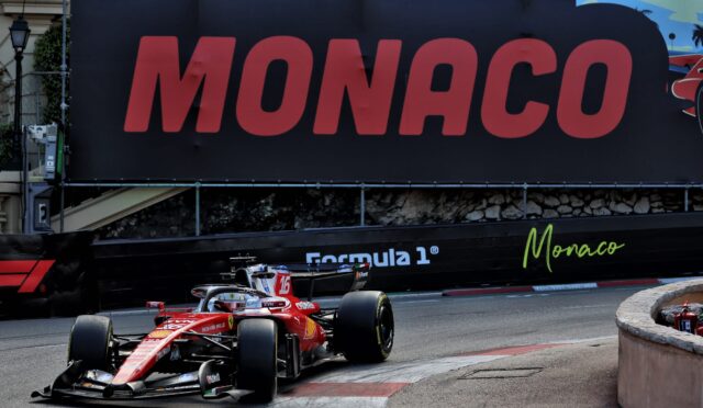 Red Formula 1 car on a tight Monaco street circuit, with a large Monaco sign on the barrier in the background.