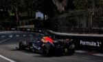 Two Formula 1 cars race along a track next to a tall iron fence and dark green trees in the background with sponsor logos on the barriers.