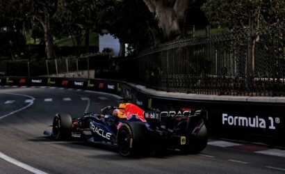 Two Formula 1 cars race along a track next to a tall iron fence and dark green trees in the background with sponsor logos on the barriers.