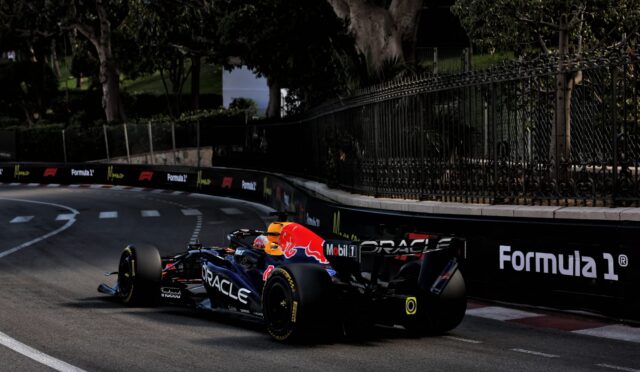 Two Formula 1 cars race along a track next to a tall iron fence and dark green trees in the background with sponsor logos on the barriers.