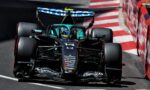 Formula One car in teal and black speeds along a race track, close to red-and-white barriers, with sponsor logos on the nose and wings nearby