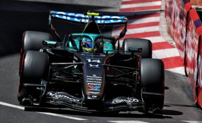 Formula One car in teal and black speeds along a race track, close to red-and-white barriers, with sponsor logos on the nose and wings nearby