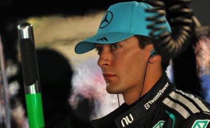 Portrait of a man in a turquoise cap with a Mercedes logo, wearing a TeamViewer racing suit and an earpiece, looking to the right.