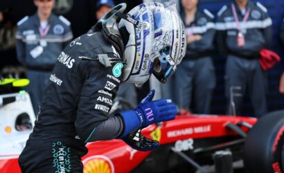 Race car driver in a dark sponsor-covered suit and blue gloves stands beside a red race car in the pit area, helmeted and ready for action.
