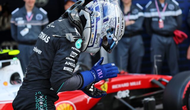 Race car driver in a dark sponsor-covered suit and blue gloves stands beside a red race car in the pit area, helmeted and ready for action.