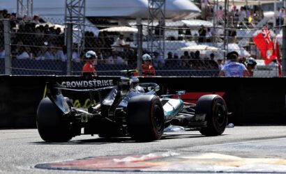 Two open-wheel Formula 1 cars on the pit lane, crowd behind a chain-link fence watching.
