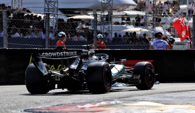 Two open-wheel Formula 1 cars on the pit lane, crowd behind a chain-link fence watching.