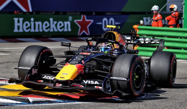 Red Bull Racing Formula 1 car on a race track near the curb, yellow nose, with Heineken ad and green barriers in the background.