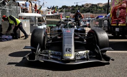 Front view of a silver Formula 1 car in the pit lane, with crew and spectators in the background beneath a busy pit area fence.