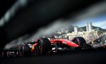 Red Formula 1 car on a Monaco street circuit with spectators and city buildings in the background, viewed through a blurred barrier in the foreground.