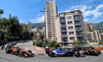 Formula 1 race cars speed along a city street circuit with spectators on balconies and tall buildings nearby.