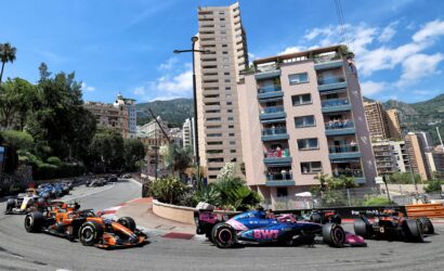 Formula 1 race cars speed along a city street circuit with spectators on balconies and tall buildings nearby.