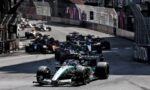 A group of Formula 1 cars race down a narrow city street circuit, lined with tall fences and advertising barriers on both sides.