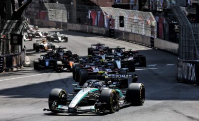 A group of Formula 1 cars race down a narrow city street circuit, lined with tall fences and advertising barriers on both sides.