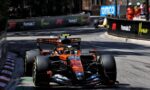 Orange Formula 1 car heading toward the camera on a city street circuit, barriers with sponsor logos lining the track on both sides.