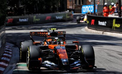 Orange Formula 1 car heading toward the camera on a city street circuit, barriers with sponsor logos lining the track on both sides.
