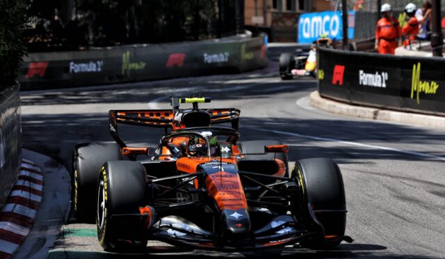 Orange Formula 1 car heading toward the camera on a city street circuit, barriers with sponsor logos lining the track on both sides.