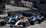 Dense pack of Formula 1 cars racing on a city street circuit with barriers along the sides and blurred spectators in the background