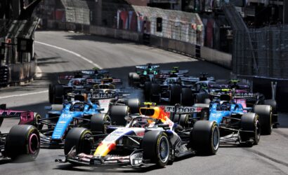 Dense pack of Formula 1 cars racing on a city street circuit with barriers along the sides and blurred spectators in the background
