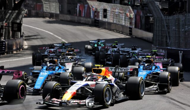 Dense pack of Formula 1 cars racing on a city street circuit with barriers along the sides and blurred spectators in the background