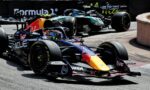 Three Formula 1 cars race closely on a track, with a Red Bull car in the foreground and others behind it on a tight bend.