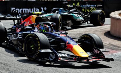 Three Formula 1 cars race closely on a track, with a Red Bull car in the foreground and others behind it on a tight bend.