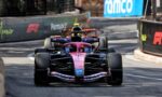 Pink Formula 1 car leading a race on a city street circuit with barrier walls on sides and other cars behind it