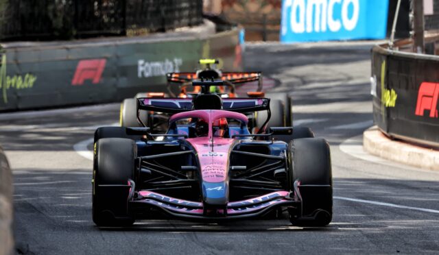 Pink Formula 1 car leading a race on a city street circuit with barrier walls on sides and other cars behind it