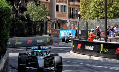 Formula 1 car in teal livery navigates a tight Monaco street circuit chicane, barriers and sponsor banners on both sides with spectators watching the action.