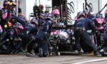 Pit crew in navy and pink uniforms perform a tire change around a race car in the pit area.