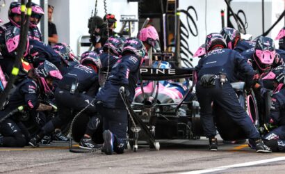 Formula 1 pit crew in navy and pink uniforms quickly servicing a pink-and-blue race car.