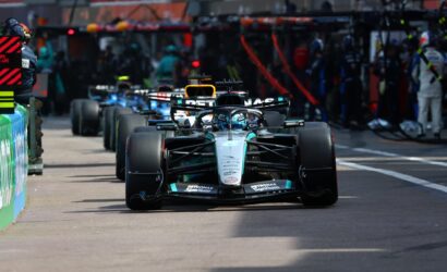 Mercedes F1 car leading a pit-lane lineup with crew and other cars in the background.