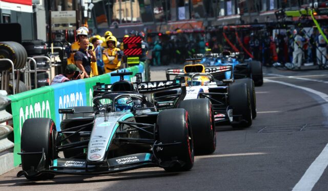 Mercedes AMG Petronas F1 car leading a line of race cars along a city street circuit, with pit crews and spectators in the background on the barriers.