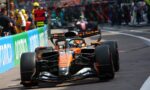 Orange Formula 1 car speeding along a city street, crowd behind barriers watching the race, front wing and wheels visible.