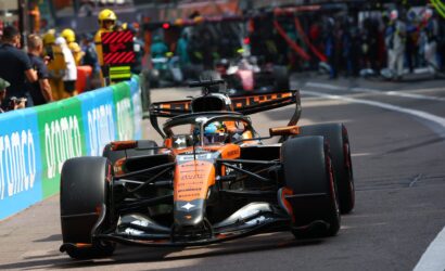 Orange Formula 1 car speeding along a city street, crowd behind barriers watching the race, front wing and wheels visible.
