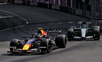 Three Formula 1 cars race on a city street circuit with barriers and sponsor banners along the wall behind them.